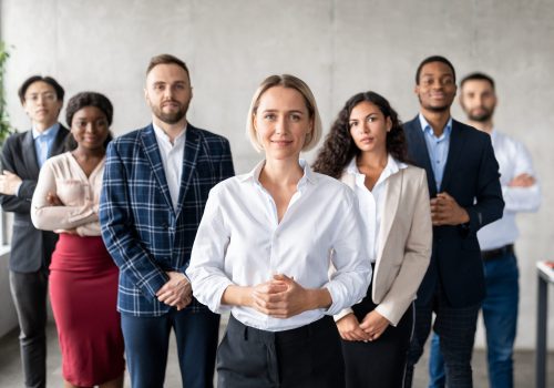 Successful Businesswoman Standing In Front Of Business Team Staff In Modern Office, Smiling To Camera. Leadership, Female Entrepreneurship And Career Growth Motivation Concept. Selective Focus